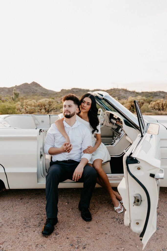 white vintage car engagement photos in the desert of Phoenix Arizona, the convertible door is open and the coupld is draped around each other sitting on the side