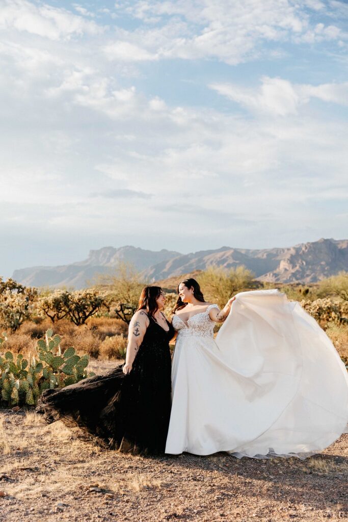 LGBTQ+ lesbian couple in wedding dresses, one in a white dress and one in a black dress, both tosing the dresses to make the train big