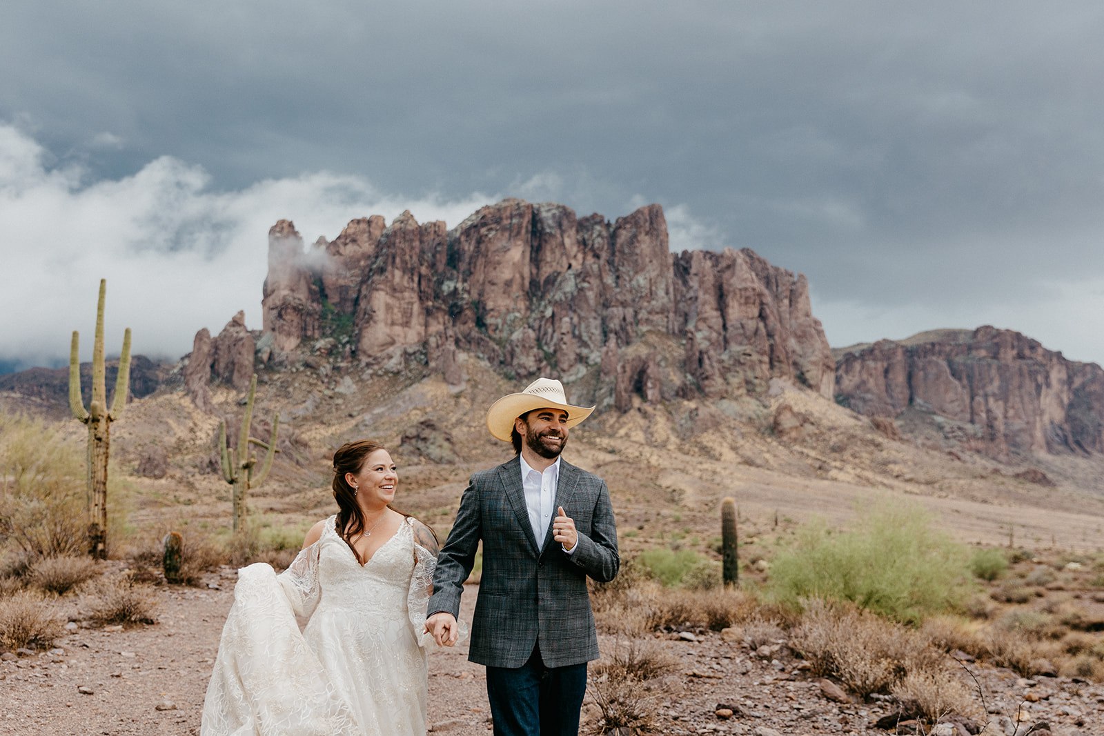 Eloping couple holding hands and running in one of the best arizona elopement locations, the iconic superstition mountains. Dark stormclouds are over the mountains