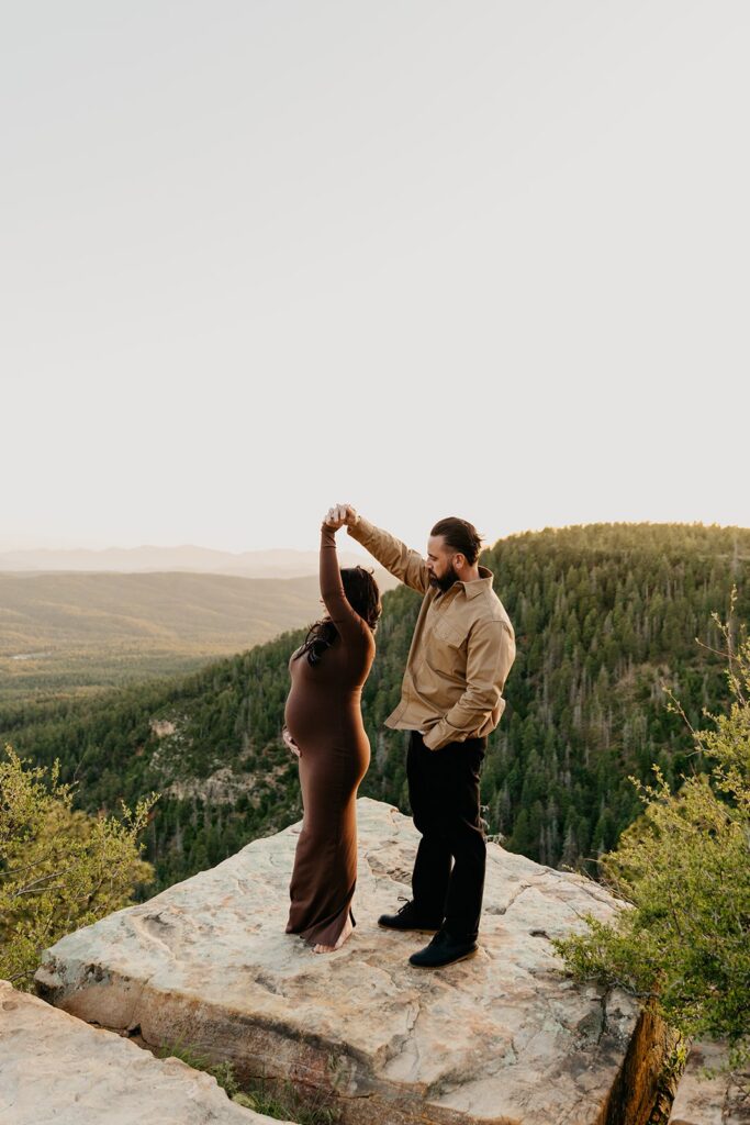 Pregnant woman getting twirled by her husband on the edge of a cliff, with pine trees and a sunset in the distance
