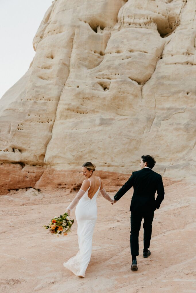 Eloping couple walking away, holding hands, towards a white stone cliff in Page, AZ